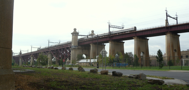 Ein Zug überquert eine Brücke mit Stützen, Strommasten mit Drähten, Schilder an Pfählen, einem Zaun, Gras, Steinen, einer Straße, Bäumen, Gebäuden und einem bewölkten Himmel im Hintergrund.
