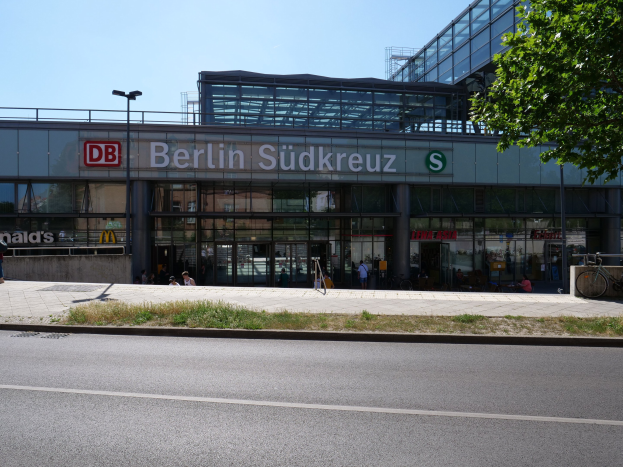 Große gläserne Berliner Südkreuz-Bahnhof mit sichtbarem Text, umgeben von Straßenmasten, Lichtern, Fahrzeugen, Fürnissen, Fahrrädern, Bäumen und einem klaren blauen Himmel.