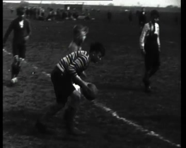 Schwarzes und weißes Foto junger Männer, die Rugby auf einem Feld spielen, einer hält den Ball.