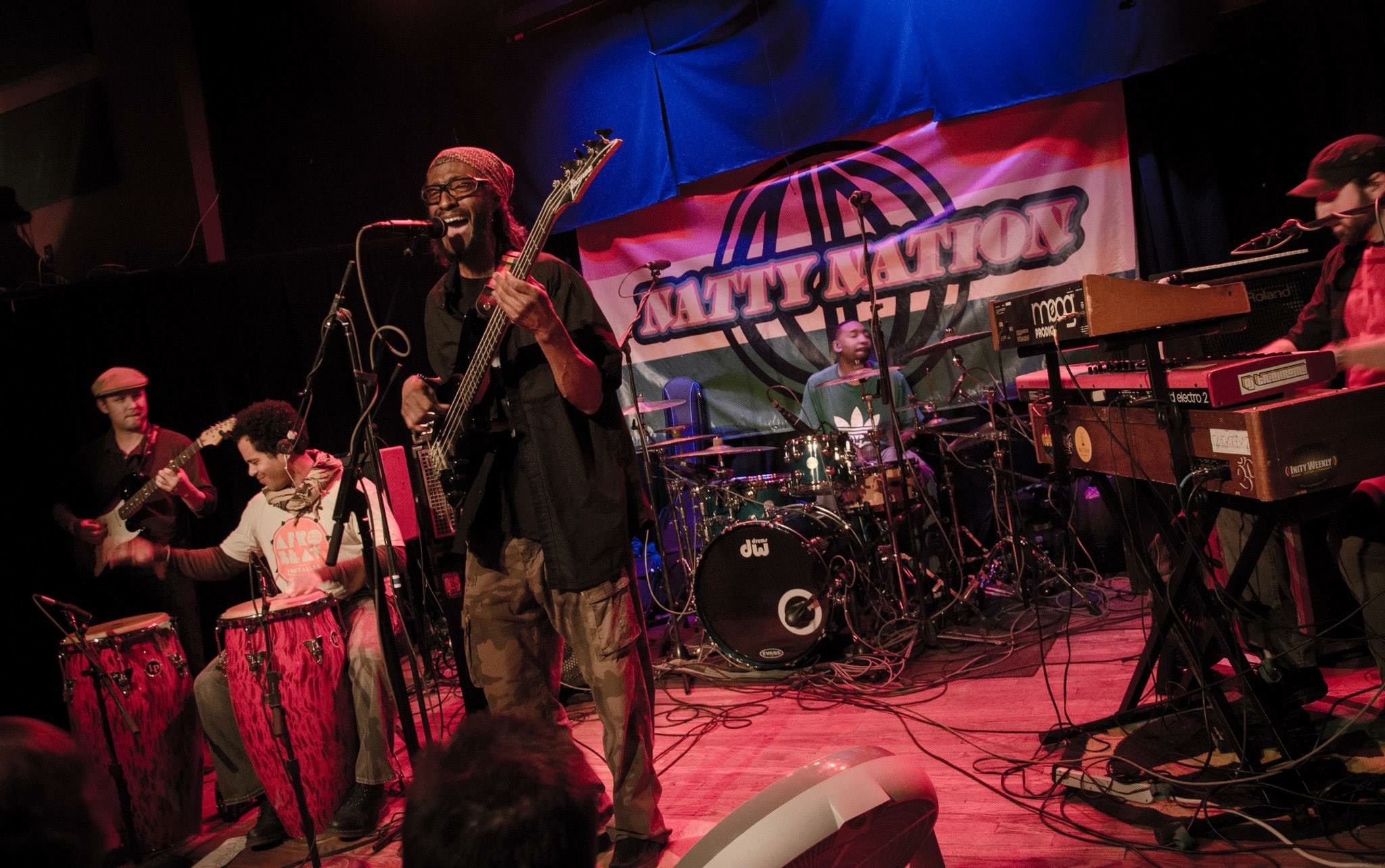 Eine Live-Band performt auf der Bühne mit Mikrofonen und Ständern, während das Publikum unten zuschaut; ein Banner mit Text ist im Hintergrund sichtbar.