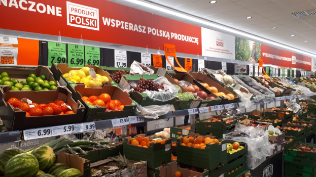 Ladeninneres mit verschiedenen Obst- und Gemüsesorten in Kartonschachteln mit Preisschildern, beleuchtet von Deckenlampen und mit informativen Tafeln an den Wänden.