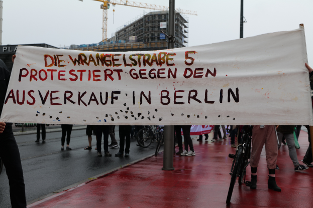 Eine Gruppe von Menschen steht auf einer Straße und hält ein Transparent mit der Aufschrift "Die Wrangellstraße 5 protestiert gegen den Ausverkauf in Berlin". Im Hintergrund sind Fahrräder, Stäbe, Gebäude und ein Kran zu sehen.