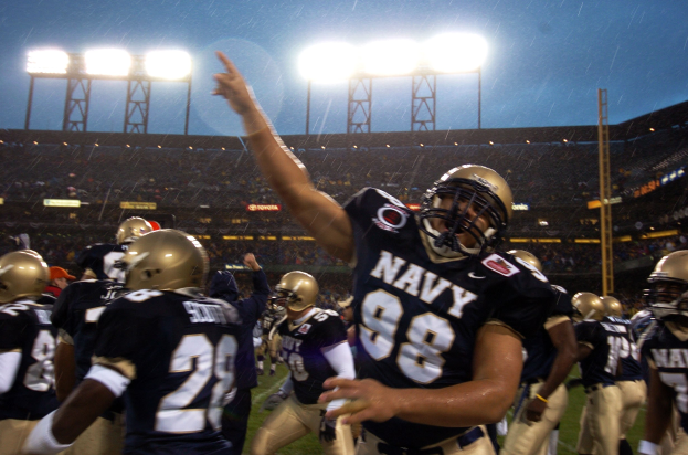 Navy-Football-Spieler feiern Touchdown auf regennasser Wiese, mit Helmen und Uniformen, Zuschauern und Stadionbeleuchtung im Hintergrund.