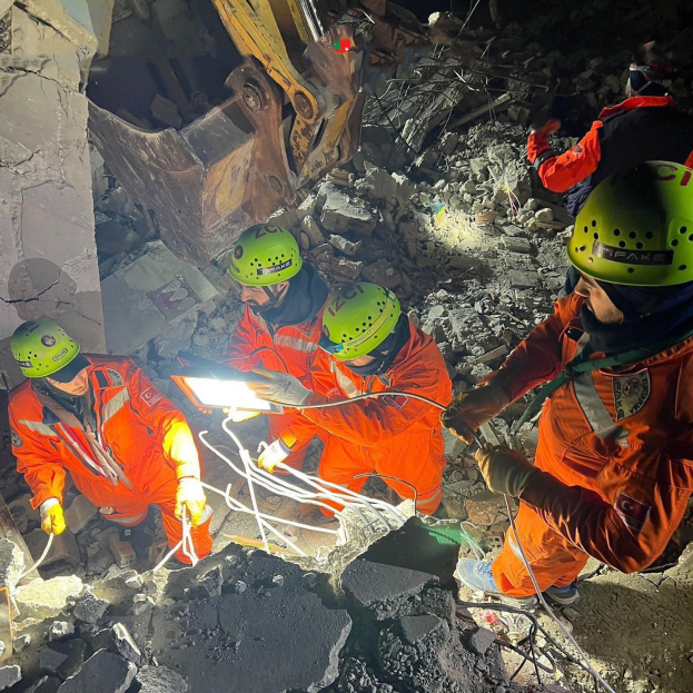 Eine Gruppe von Menschen in orangefarbenen Uniformen und grünen Helmen bei der Arbeit an Schutt von einem eingestürzten Gebäude mit einem Kran im Hintergrund, die Rettungsgeräte und -materialien halten.