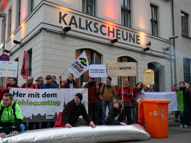 Eine Gruppe von Menschen mit Protestschildern und Plakaten vor einem Gebäude, mit zwei Personen im Vordergrund und einem Müllcontainer rechts daneben, vor einem Hintergrund von Gebäuden mit Fenstern und Schildern in Deutschland.