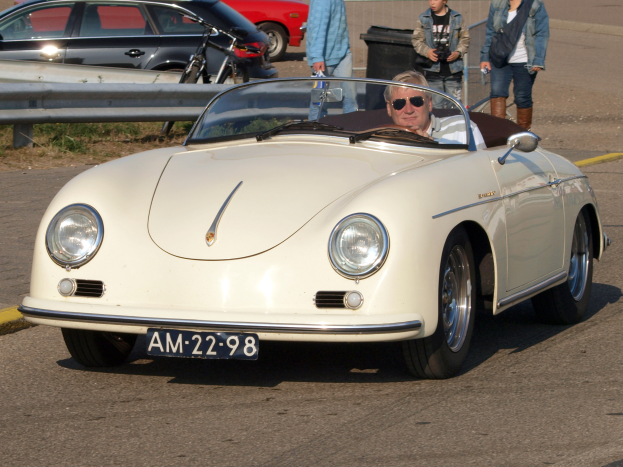 Ein Mann fährt einen weißen Porsche 356 Speedster die Straße entlang, umgeben von Menschen in verschiedenen farbigen Kleidern, mit einem Geländer und einem schwarzen Müllcontainer an der Seite und weiteren Autos und einem Gebäude im Hintergrund.