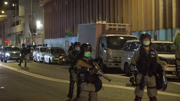 Polizeibeamte in Helmen, Handschuhen und Gesichtsmasken stehen auf einer Straße bei Nacht, einige halten Waffen, mit Fahrzeugen, Laternenpfählen, Schildern und Gebäuden im Hintergrund.