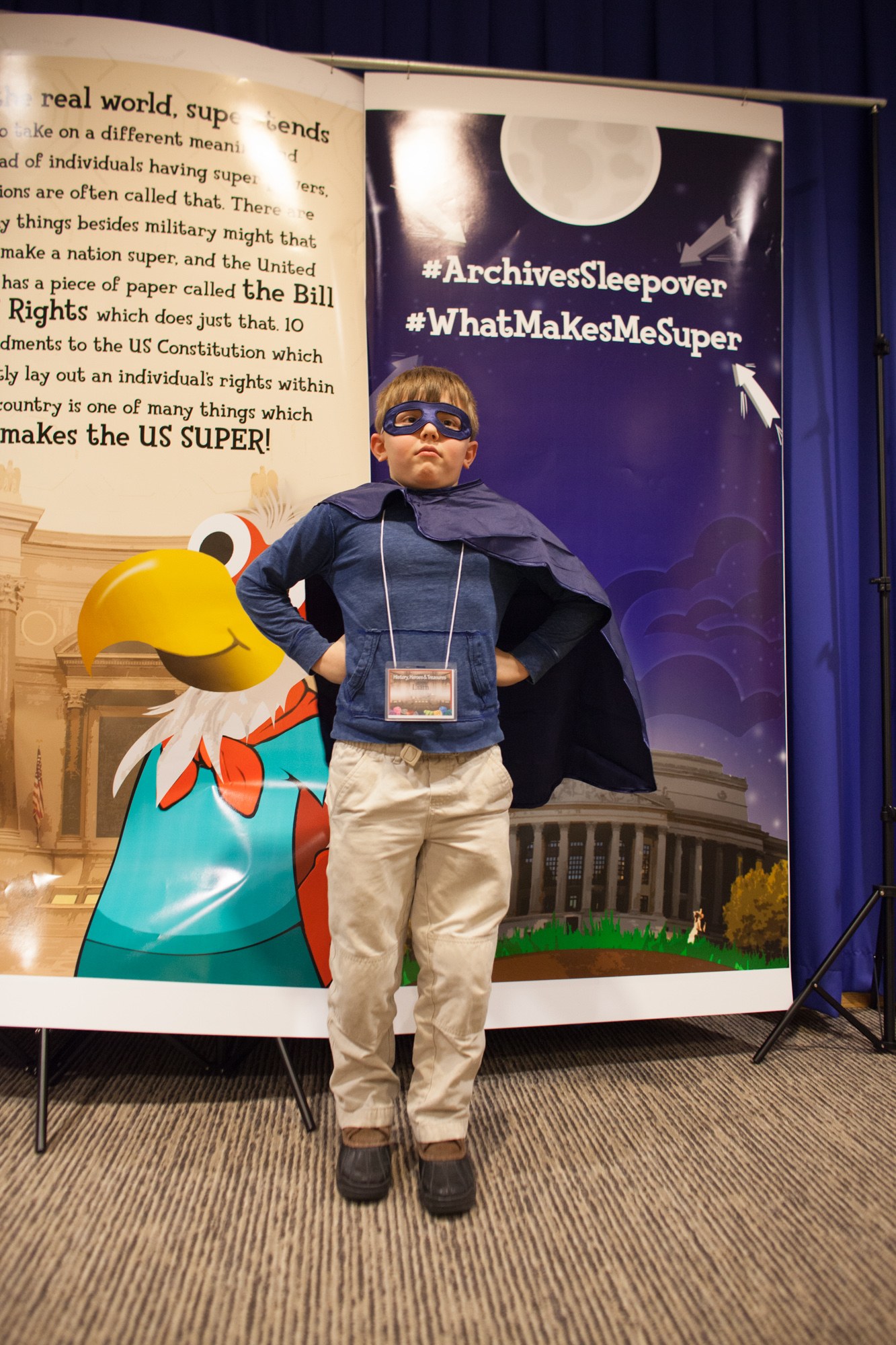 Ein junger Junge in einem Superheldenkostüm steht vor einem Banner mit Text und einem Vogelbild, mit einem Vorhang im Hintergrund.