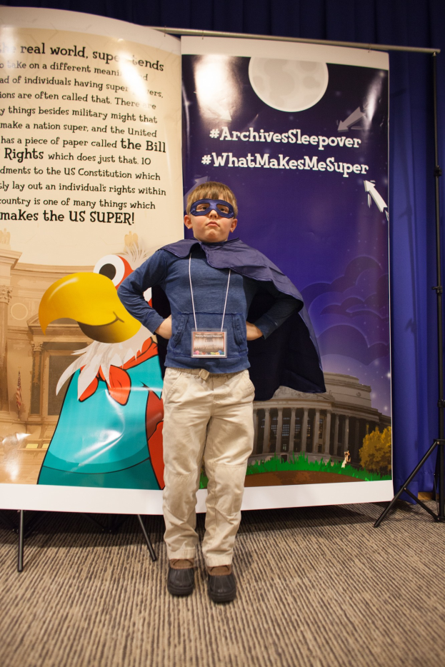 Ein junger Junge in einem Superheldenkostüm steht vor einem Banner mit Text und einem Vogelbild, mit einem Vorhang im Hintergrund.