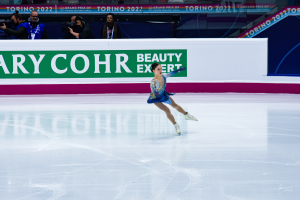 Eine Frau in einem blauen Kleid fährt auf einem Eisstadion, während eine Menge mit Kameras zusieht, mit einem Schild im Hintergrund, auf dem 'Tessa Virtue und Scott Moir' steht.