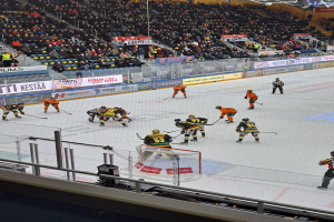 Eine Gruppe von Menschen, die Hockey auf einem Eisplatz spielen, umgeben von einem Zaun, mit Zuschauern im Hintergrund und beleuchtet von Scheinwerfern an der Decke.