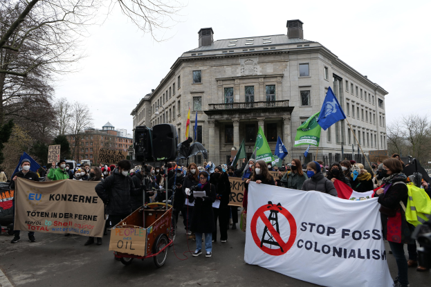 Große Gruppe von Menschen bei einer Demonstration gegen fossile Brennstoffe, die Transparente und Fahnen tragen, mit einem Fahrzeug im Vordergrund und Gebäuden, Bäumen und einem klaren blauen Himmel im Hintergrund.