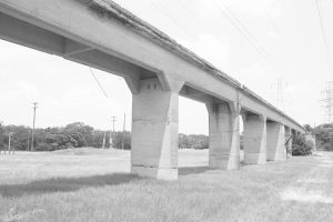 Eine Betonbrücke auf einer grünen Wiese mit einem großen elektrischen Pfahl und Kabeln darüber, umgeben von Bäumen und zusätzlichen elektrischen Pfählen im Hintergrund.