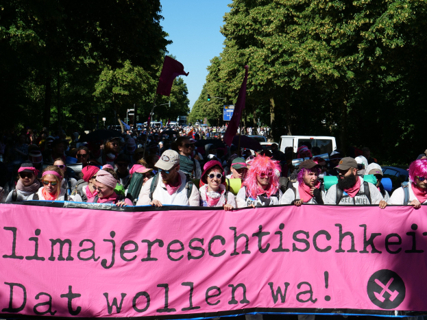 Eine Gruppe von Menschen marschiert die Straße entlang, hält ein pinkes Banner mit der Aufschrift "Women's March in Berlin" hoch, einige tragen Mützen und Schutzbrillen und andere halten Fahnen, Bäume und Fahrzeuge im Hintergrund.