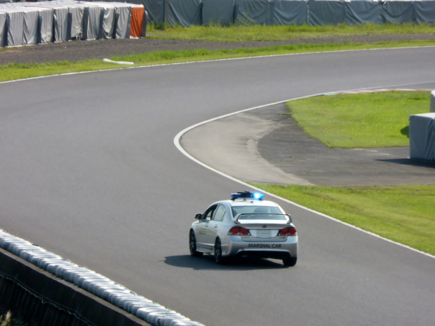 Polizeiauto auf einer Rennstrecke mit Gras auf beiden Seiten, mit Menschen, Zelten und anderen Gegenständen im Hintergrund.