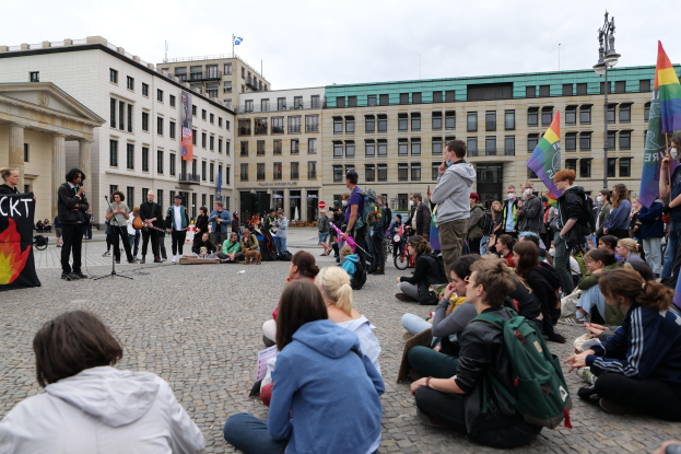 Eine Gruppe von Menschen, die auf dem Boden vor einer Menge sitzen, die Fahnen und Transparente hält, während einer Anti-Schwulen-Demonstration in Berlin, mit einer Statue, Gebäuden und einem bewölkten Himmel im Hintergrund.
