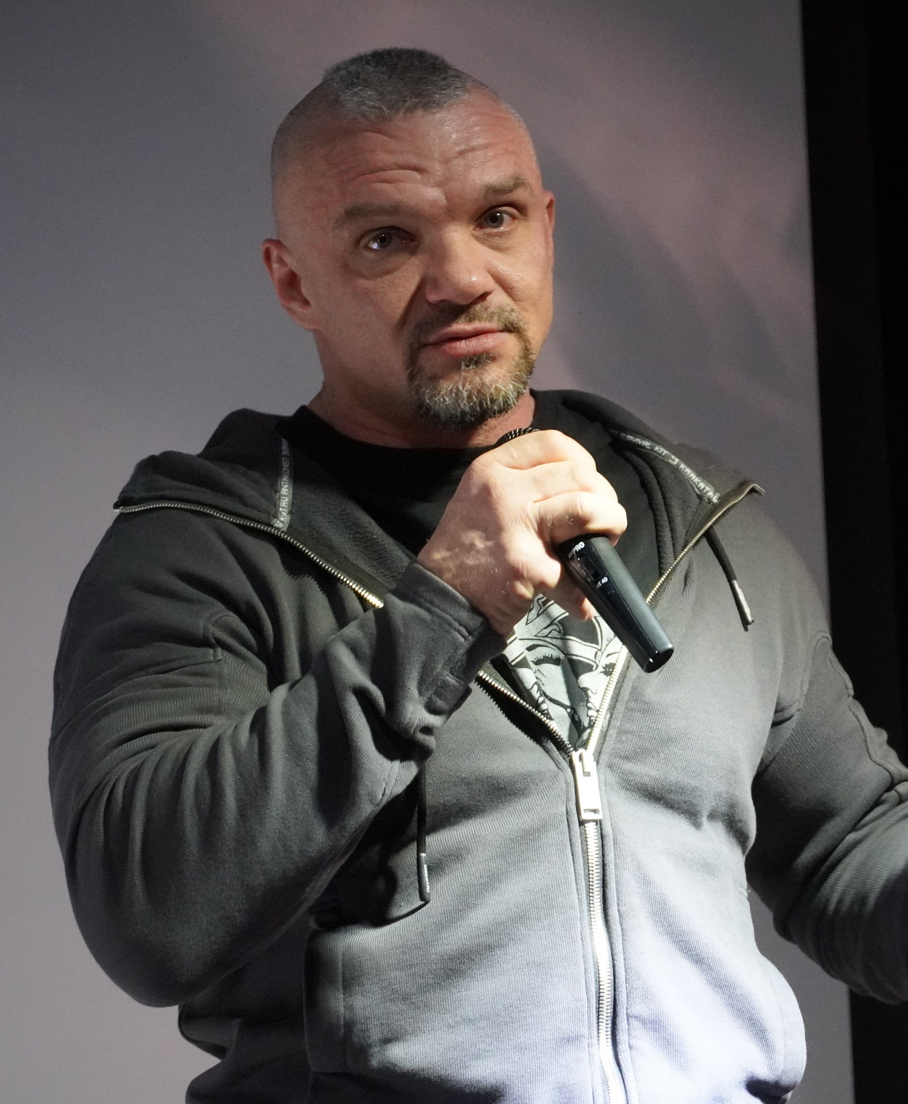 Ein Mann in einer schwarzen Kapuzenjacke und einem schwarzen Anzug, der ein weißes Hemd trägt, hält ein Mikrofon und hat zurückgekämmtes Haar und einen selbstbewussten Gesichtsausdruck, während er vor einer Wand steht.