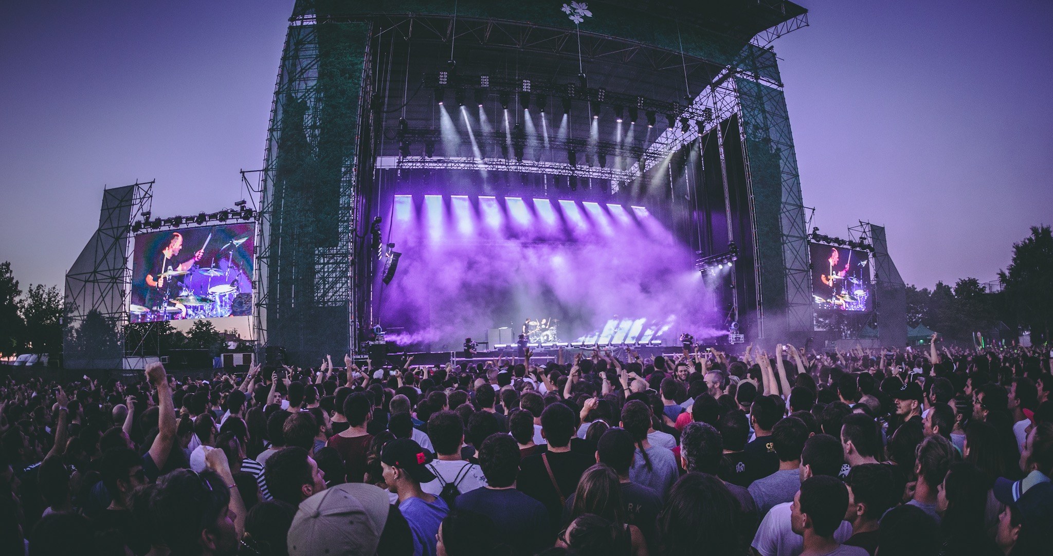 Eine große Menschenmenge bei einem Musikfestival schaut auf eine beleuchtete Bühne mit Instrumenten und Bildschirmen, die vor einem Gebäude, Bäumen und einem blauen Himmel aufgebaut sind.