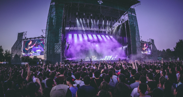 Eine große Menschenmenge bei einem Musikfestival schaut auf eine beleuchtete Bühne mit Instrumenten und Bildschirmen, die vor einem Gebäude, Bäumen und einem blauen Himmel aufgebaut sind.