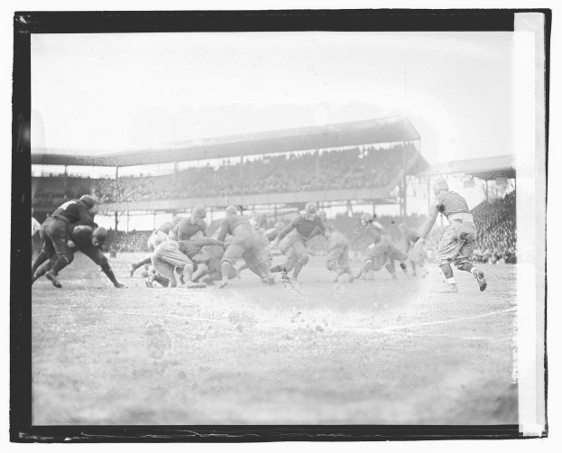 Eine Gruppe von Männern, die Fußball auf einem Feld mit einem Stadion im Hintergrund und einem klaren Himmel spielen, dargestellt in Schwarz-Weiß.