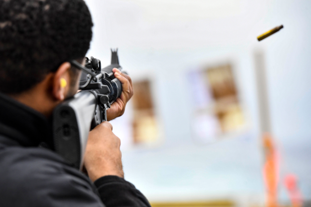 Ein Mann in einer schwarzen Jacke zielt mit einem Gewehr auf ein Ziel, mit einer Kugel in der Luft und einem unscharfen Hintergrund.