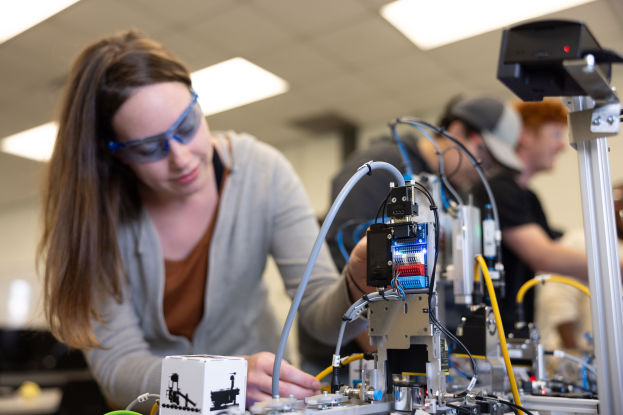 Eine Frau in Schutzbrille arbeitet an einem Roboter in einem Labor umgeben von anderen, mit verschiedenen Gegenständen auf dem Tisch.