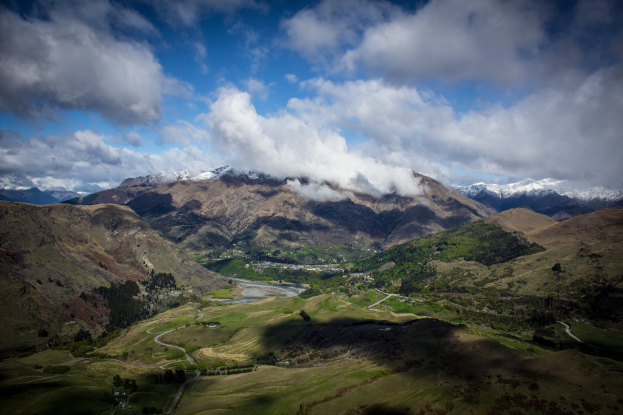 Eine Panoramansicht vom Gipfel eines Berges in Queenstown, Neuseeland, zeigt saftig grünes Gras, verstreute Bäume und eine gewundene Straße unter einem Himmel voller weißer, flauschiger Wolken.