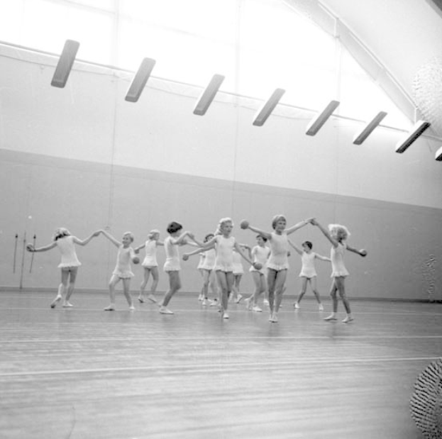 Schwarz-weißes Bild von jungen Mädchen in einem Tanzstudio, die sich an den Händen halten und tanzen, mit einem Ball auf der rechten Seite und Lampen an der Decke.