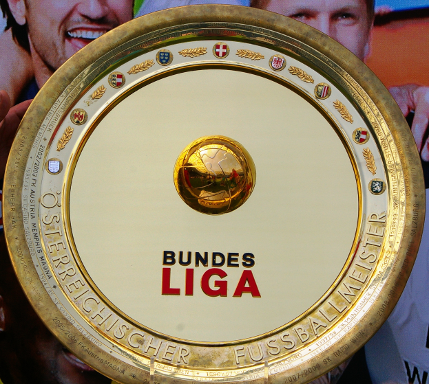 Ein Mann hält einen goldenen Schild mit 'Bundesliga' darauf, mit einem Banner im Hintergrund mit drei Personen.