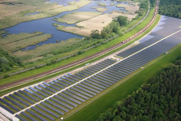 Luftaufnahme einer Solar-Farm mit Solar-Panelen auf einer Wiese umgeben von Bäumen und Wasser, mit einem Zug auf einer nahen Bahnstrecke.
