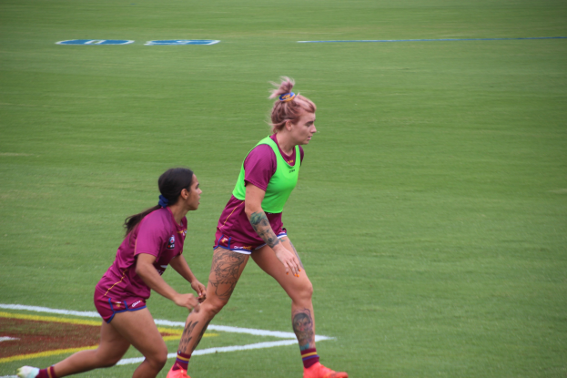 Zwei Frauen beim Fußballspielen auf einem Feld, eine in einem grünen T-Shirt und Shorts, die andere in einem pinken Kleid, beide in Sportkleidung und sichtbare Tattoos an Händen und Füßen.