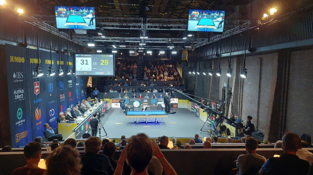 Eine große Menschenmenge sitzt vor einer Bühne mit einem Tischtennis-Tisch in der Mitte, einer Wand auf der rechten Seite und einem Publikum im Hintergrund unter Deckenlampen und Fernsehern, die als die 2019 Jumbo-Weltmeisterschaft gekennzeichnet sind.