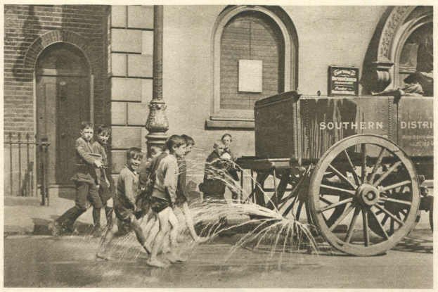 Schwarz-weißes Bild von Kindern, die mit einem Feuerhydranten vor einem Gebäude mit Fenstern, einer Tür, einem Mast und einem Zaun spielen, mit einem Wagen im Hintergrund.