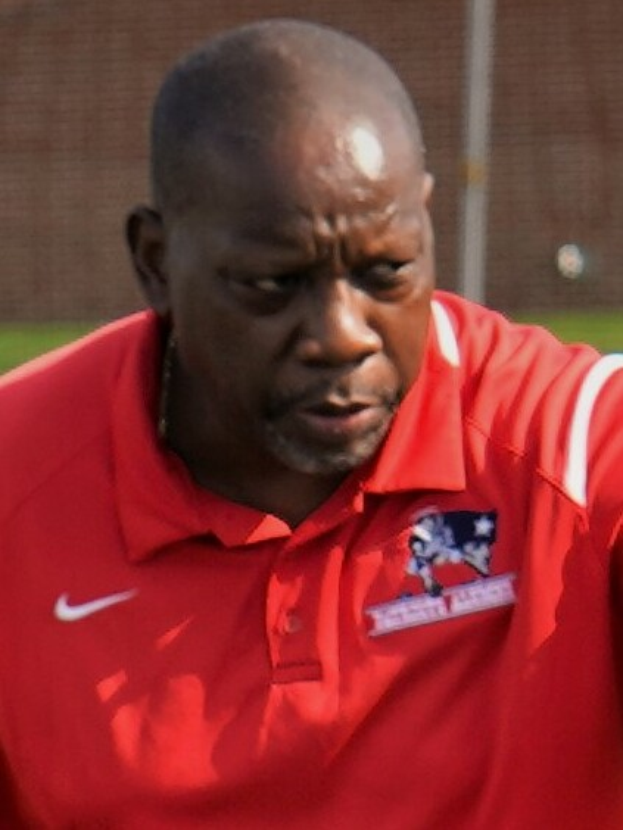 Ein Mann in rotem T-Shirt steht auf einem Feld und hat die Hände in die Hüften gestützt, er sieht ernst aus, der Hintergrund ist unscharf.