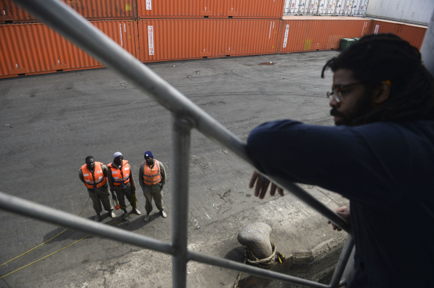 Ein Mann in legerer Kleidung steht auf einem Metallgelönder in der Nähe einer Gruppe von Menschen mit orangen Jacken und Kappen, mit Containern und einer Wand im Hintergrund am Hafen von Los Angeles.