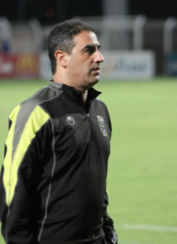 Ein Mann in einer schwarzen und gelben Jacke steht auf einem Fußballfeld, wahrscheinlich der Trainer, mit einem unscharfen Hintergrund.