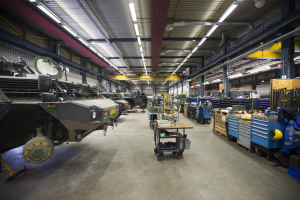 Ein großes Lager voller Maschinen, einschließlich Fahrzeugen auf der linken Seite, Wagen mit Gegenständen, Holzboxen und anderen Gegenständen auf der rechten Seite, mit Lampen oben.