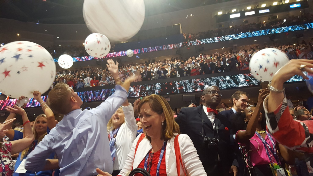 Eine Menschenmenge steht vor einem großen Publikum, einige halten Luftballons, andere sitzen im Hintergrund unter beleuchteten Lichtern, wahrscheinlich feiern sie die Republican National Convention.