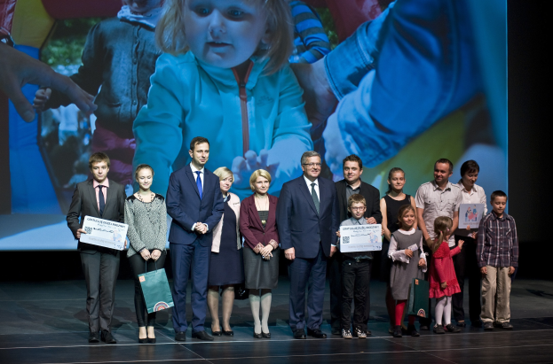 Eine Gruppe von Menschen auf einer Bühne mit Taschen und Urkunden, im Hintergrund ein Bildschirm mit einem Bild eines Kindes und anderen Menschen.