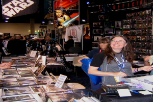 Eine Frau posiert an einem Kassenautomaten in einem belebten Geschäft, das mit Bildern, Büchern und ausliegenden Plakaten gefüllt ist, im Hintergrund kaufen Kunden ein.