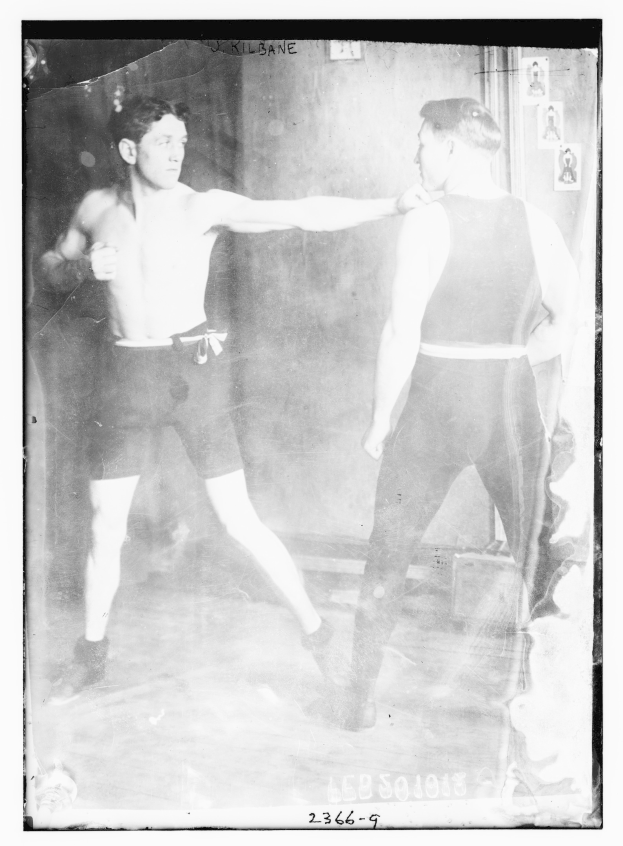 Schwarüß-weißes Foto von zwei Boxern in einem Boxring mit Plakaten an der Hintergrundwand und Text unten.