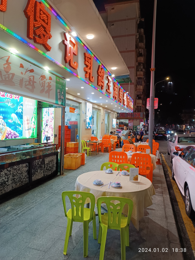 Outdoor restaurant seating at night with tables set with utensils, a few people seated, and buildings illuminated by street lights.