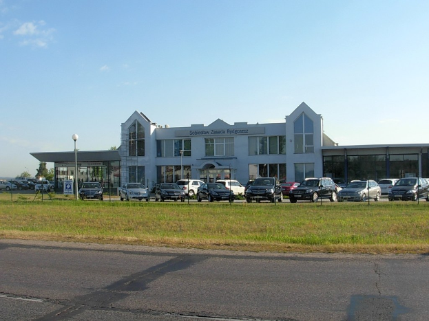 Autohaus mit parkenden Autos, umgeben von Gebäuden, Straßeninfrastruktur, fahrenden Fahrzeugen, Bäumen und einem bewölkten Himmel.