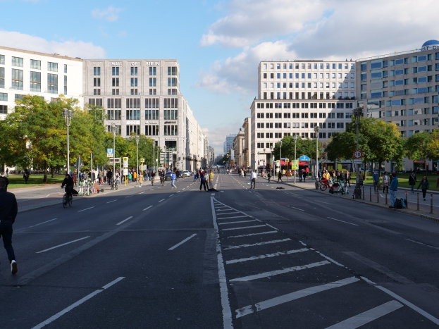 Eine belebte Stadtstraße in Berlin mit Fußgängern, Radfahrern, hohen Gebäuden, Bäumen, Laternenmasten und Schildern unter einem bewölkten Himmel.