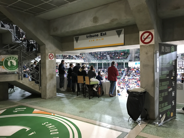 Eine Gruppe von Menschen steht vor einer sitzenden Menge in einem Stadion während eines Boston Celtics vs New York Jets-Spiels, mit sichtbaren Schildern, einem Müllkorb, einer Texttafel, einer Treppe mit Gelöndern und Deckenlampen.