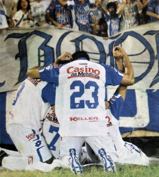 Gruppe von Fussballspielern, die auf dem Boden vor einer jubelnden Menge feiern, mit einem teilweise sichtbaren Banner im Hintergrund.