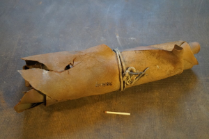 Ein brauner Papiertüte in Form einer Granate mit Henkel, verschnürt mit einem Seil und einer Zigarette daneben auf einem Tisch.