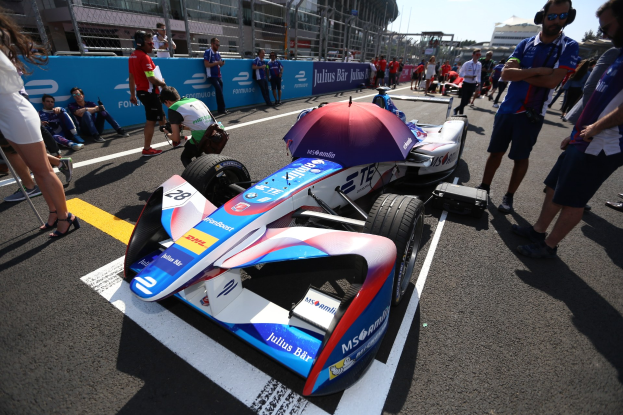 Formula E-Rennwagen mit weißer, blauer, roter und schwarzer Lackierung auf einer Strecke geparkt, umgeben von Menschen, mit einem Schirm im Vordergrund und Renninfrastruktur im Hintergrund unter einem klaren Himmel.