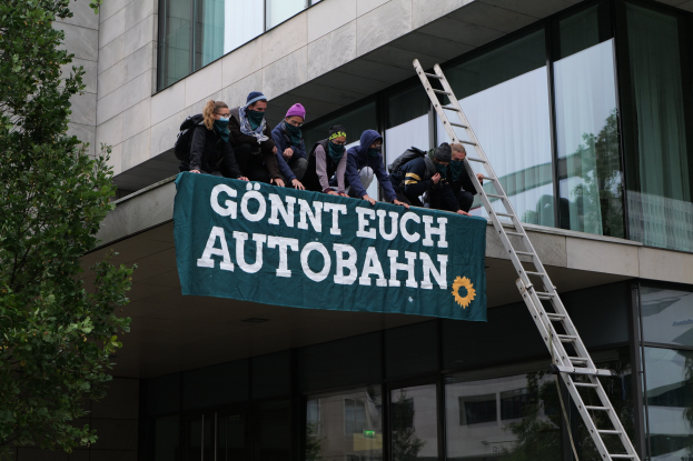 Eine Gruppe von Menschen auf dem Dach eines Gebäudes mit einer Fahne, einem Baum auf der linken Seite und Glasfenstern im Hintergrund, wahrscheinlich protestieren sie für die deutsche Regierung.