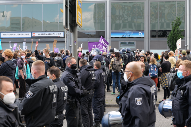 Eine große Gruppe von Menschen steht vor einem Gebäude, einige halten Schilder und tragen Helme, mit einem Schildständer im Vordergrund und einem Baum im Hintergrund, scheinbar protestierend gegen eine deutsche Regierungspolitik.
